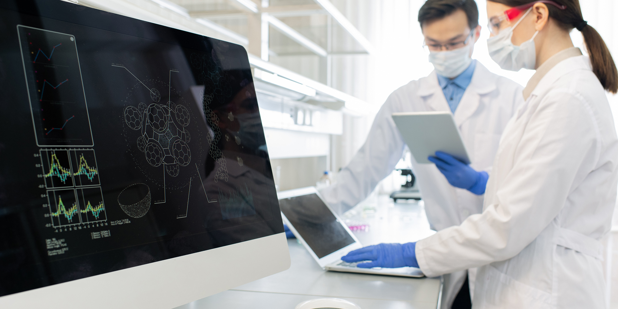 researchers wearing mask and gloves collaborating in laboratory beside computer screen